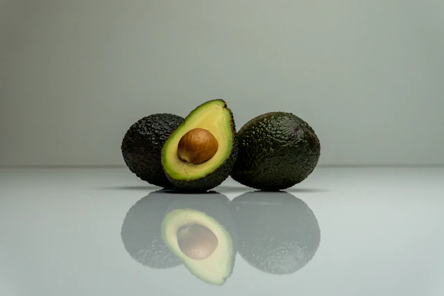 A fresh avocado half with two whole avocados on a pale reflective surface.
