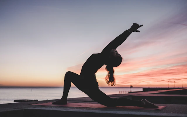 A person stretching on a mat beside the water at sunrise.
